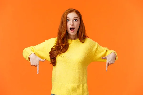 Regarde vite. Incroyable et fasciné fille gingembre mignon, étudiant rousse en pull jaune, haletant bouche ouverte de surprise et d'émerveillement, pointant les doigts vers le bas, look impressionné, fond orange — Photo