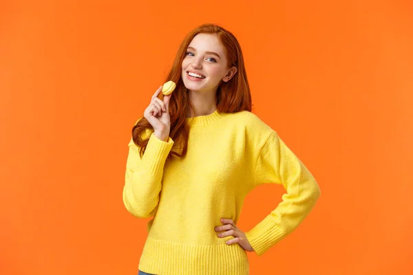 Retrato en la cintura encantadora pelirroja sosteniendo sabroso macaron amarillo cerca de la boca y sonriendo alegremente, como comer dulces, comprar postre para la cita romántica de San Valentín, posando fondo naranja alegre — Foto de Stock