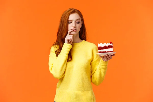 饮食，健康的生活方式和垃圾食品的概念。 可爱的红头发女人忍不住咬好吃的蛋糕，把甜点放在盘子里，咬着嘴唇，想着里面有多少卡路里，橙色的背景 — 图库照片