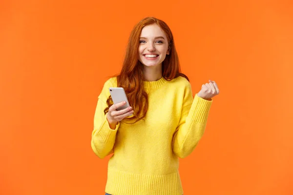 Mutlu kız seninle iyi haberleri paylaşıyor. Neşeli, kızıl saçlı, elinde akıllı bir telefon olan güzel bir kadın zafer ya da kutlama için yumruk tokuşturuyor, oyunu kazanıyor, hoşlandığı adamdan mesaj var, turuncu arka plan.