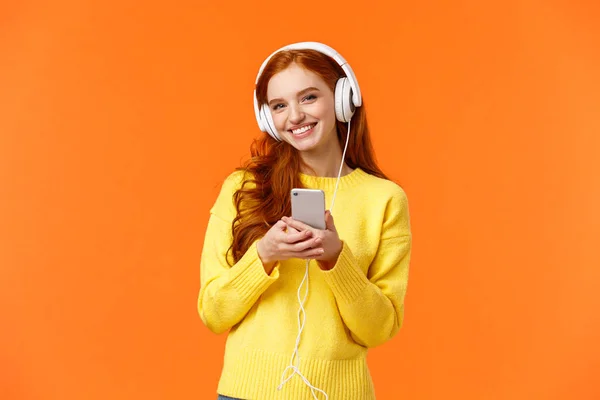 Concepto de tecnología, compras y personas. Chica pelirroja hipster lindo moderno con auriculares blancos, la celebración de teléfono inteligente, sonriendo despreocupado, disfrutar de buen sonido de la música, de pie fondo naranja — Foto de Stock