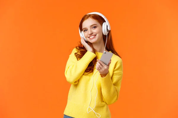 Περιμένετε-up πορτρέτο χαρούμενα όμορφη νεαρή γυναίκα με κόκκινα μαλλιά ακούστε μουσική στα ακουστικά, γείρετε το κεφάλι και χαμογελώντας ικανοποιημένοι, κρατώντας smartphone, επιλέξτε τραγούδι για την προσφορά ήρεμη βραδιά, πορτοκαλί φόντο — Φωτογραφία Αρχείου