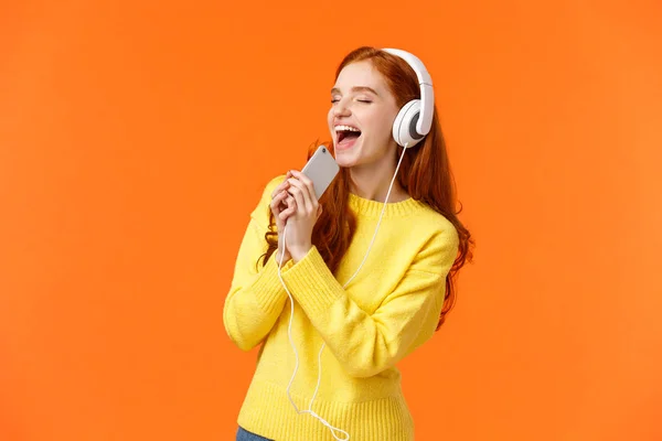 Girl carried away while listen favorite track. Pretty redhead female enjoy music in new headphones parents bought for birthday, holding smartphone close eyes and singing in microphone as if karaoke