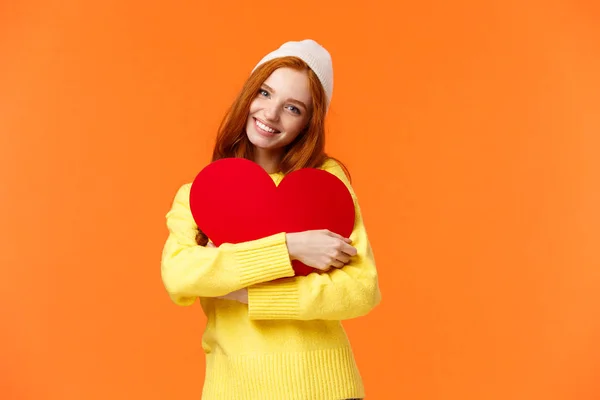 Mujer pelirroja tierna y romántica de ensueño en sombrero de invierno, suéter, abrazo grande lindo signo de corazón rojo como símbolo de amor, sonriendo alegremente, celebrando el día de San Valentín, fondo naranja —  Fotos de Stock