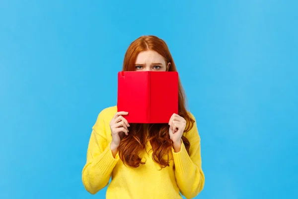 Upset gloomy and uneasy cute pouting redhead girl, frowning dont want speak to you, hiding face behind red notebook, frowning and staring offended, sulking over blue background, bad mood — Stock Photo, Image