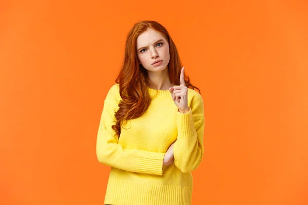 Redhead girl express disapproval as shaking her finger at someone with disappointed angry face, frowning give warning, prohibit unacceptable behaviour, standing displeased orange background