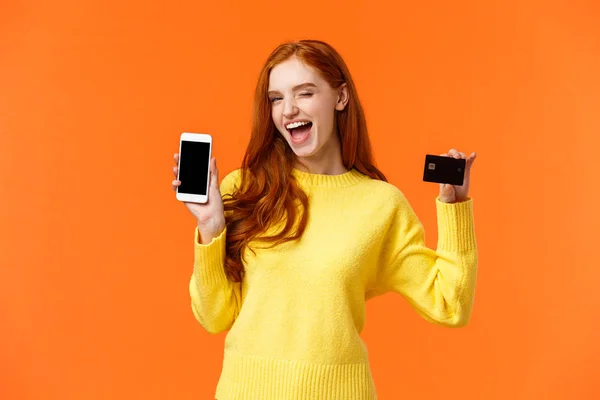 Shopping, pubblicità e mobile addication concept. Attraente ragazza dai capelli rossi sfacciati che mostra smartphone e carta di credito, occhiolino e sorriso fotocamera, promuovere online banking, deposito o metodo di pagamento — Foto Stock