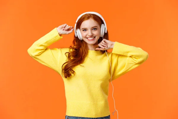 Feliz e despreocupado animado ruiva hipster menina como seus novos fones de ouvido, dançando com as mãos levantadas e sorrindo desfrutar de ouvir música sobre fundo laranja, aumentar o humor com música favorita — Fotografia de Stock