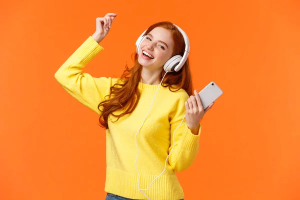 Carefree feliz ruiva adolescente receber novos fones de ouvido para presente de Natal, dança levantar a mão enquanto ouve música favorita, segurando smartphone, colocar faixa incrível, fundo laranja — Fotografia de Stock