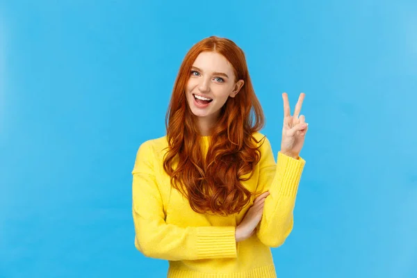 Alegre saída e feliz carismático ruiva fêmea em camisola amarela, mostrando sinal de paz, número dois ou duas vezes e sorrindo, fazendo compra, encomendar queijo duplo, fundo azul — Fotografia de Stock