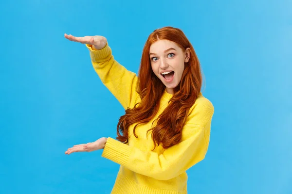 Emocionada chica pelirroja alegre y atractiva sonriendo fascinada y divertida, mostrando un objeto grande y grande, dando forma al tamaño con las manos levantadas sobre el fondo azul, de pie alegre, impresionada —  Fotos de Stock