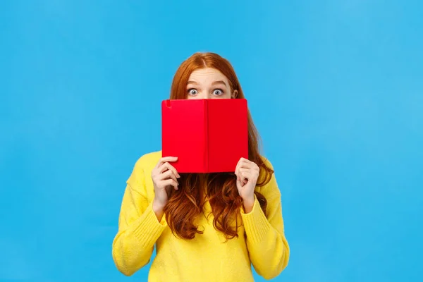 Shy and cute redhead girl like reading books and writing in diarty, hiding face behind red cool notebook, stare curious and surprised with popped eyes, standing blue background modest — Stock Photo, Image