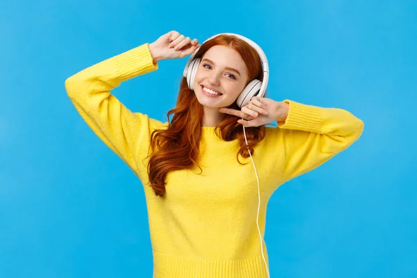 Technology, e-shopping and people concept. Modern good-looking redhead female in yellow sweater, dancing carefree, listen favorite singer music in headphones, smiling camera, blue background — Stock Photo, Image