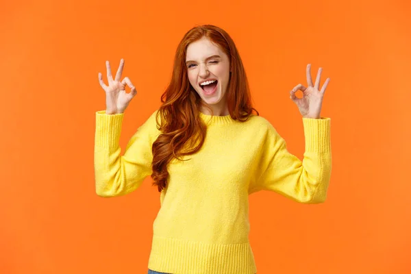 Fröhlich und positiv, enthusiastische rothaarige Frau im gelben Pullover zwinkert zustimmend, fröhlich lächelnd, gibt Erlaubnis, positive Antwort, bewertet guten Film, nickt zustimmend, empfehle, orangefarbener Hintergrund — Stockfoto