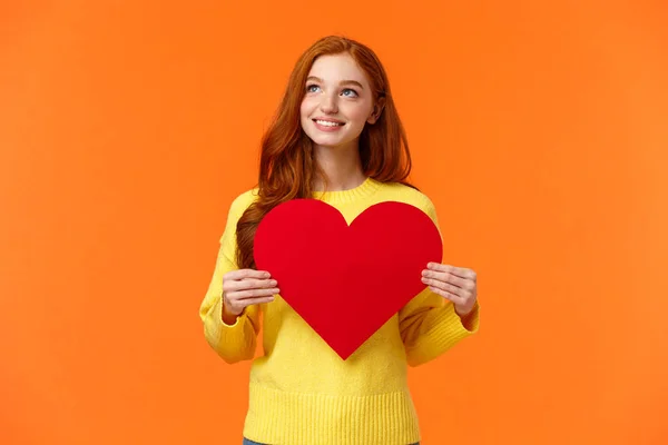 Dreamy cute redhead girl waiting for her prince, planning perfect velantines day romantic date, holding large heart sign and looking upper left corner with tender happy smile, orange background — Stock Photo, Image