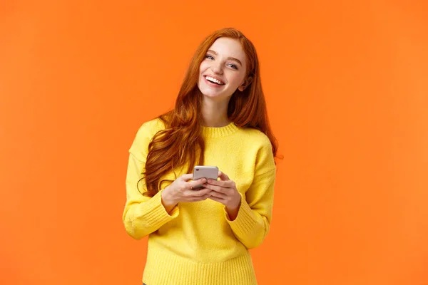 Dziewczyna szuka prezentów na wakacje online. Beztroska, szczęśliwa ruda studentka korzystająca z aplikacji na smartfona, śmiejąca się radośnie, trzymająca telefon komórkowy, czatująca lub przeglądająca portale społecznościowe, pomarańczowe tło — Zdjęcie stockowe