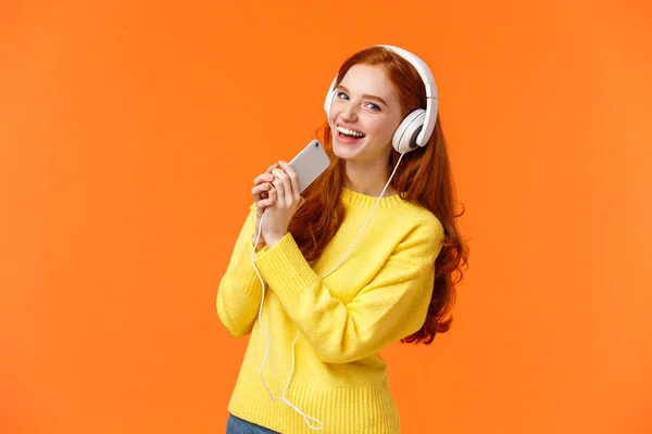 Mulher muito ruiva caucasiana gosta de ouvir música, jogando aplicativo de karaoke no smartphone com fones de ouvido, olhando a câmera com alegria, segure o telefone celular como microfone, fundo laranja — Fotografia de Stock