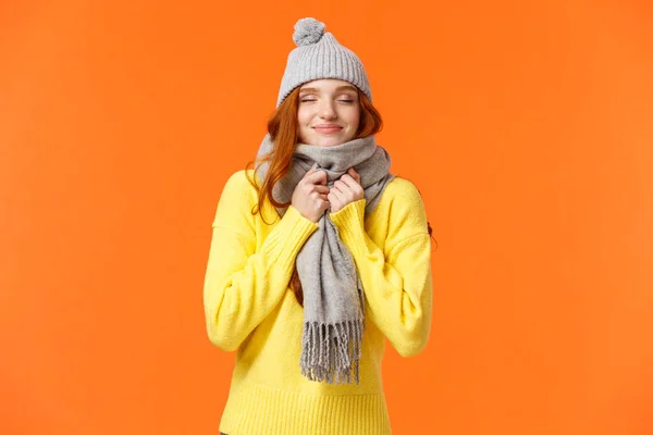 Mujer pelirroja linda y feliz caminando a lo largo de las vacaciones de invierno mercado justo, cerrar los ojos y sonreír como oliendo algo delicioso, con sombrero gris cálido y bufanda, de pie fondo naranja — Foto de Stock