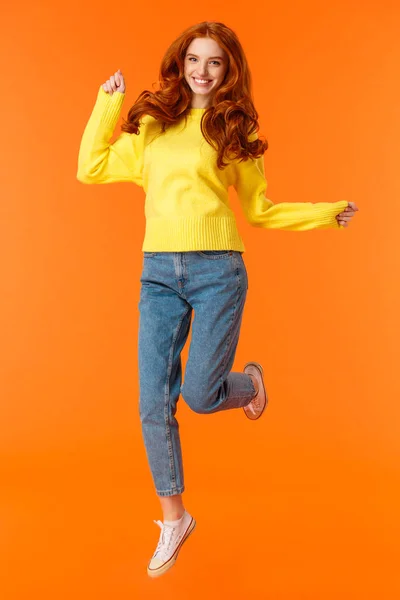 Vertical full-length retrato alegre ruiva animado menina pulando e se sentindo feliz, sorrindo olhando câmera despreocupada, não pode esperar presentes abertos no inverno festa de férias, fundo laranja — Fotografia de Stock