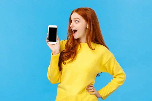 哇哦，看看这个应用程序。 兴奋而兴奋的红头发女孩看着智能手机的显示屏，展示手机屏幕、购物网站或应用程序，站在蓝色的背景上 — 图库照片