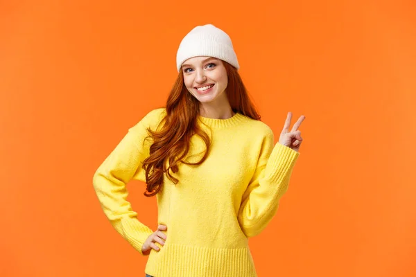 Menina moderna hipster jogando bolas de neve com amigos durante as férias de inverno, mostrando sinal de paz como posando foto perto boneco de neve, vestindo gorro branco e suéter sobre fundo laranja — Fotografia de Stock