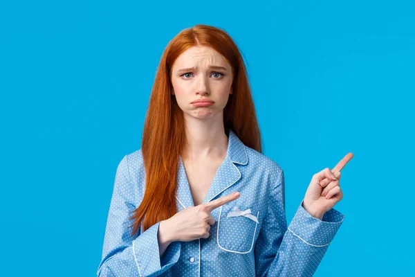 Regret, bad mood and advertising concept. Sad unhappy redhead girl in nightwear, sulking and frowning express disappointment or jealousy, pointing fingers right, standing blue background