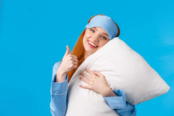 Saying yes to good healthy sleep. Cheerful pretty redhead woman in sleep mask and nightwear, hugging soft pillow showing thumbs-up in approval, like gesture, standing pleased blue background — Stock Photo, Image