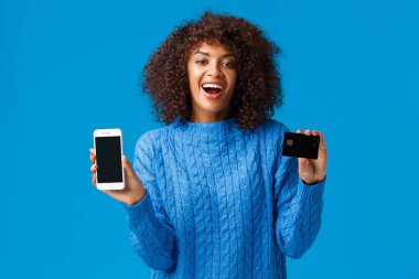 Mutlu ve heyecanlı, büyüleyici Afro-Amerikan kadın alışveriş hastası online hediyeler alıyor, başvuru, alışveriş uygulaması ve kredi kartı gösteriyor, gülümsüyor ve gülüyor, mavi arka planda duruyor.