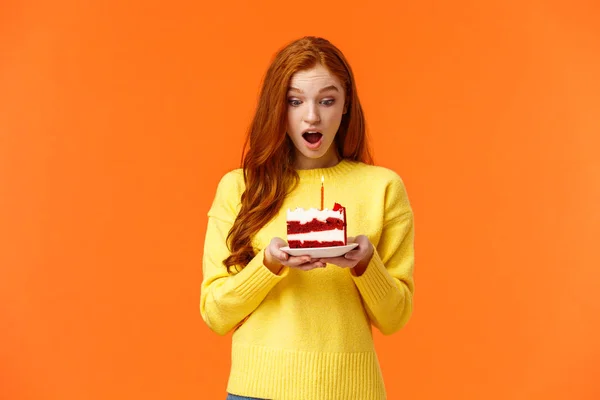 Дівчина робить бажання, отримує торт на день народження з однією свічкою, святкує б-день, отримує несподіване відчуття задоволення і вдячності, дивиться на десерт з захопленням і щасливим виразом, помаранчевий фон — стокове фото