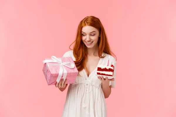 Curiosa y feliz, alegre chica pelirroja atractiva en vestido blanco, celebrando cumpleaños, recibir regalo lindo, mirando a regalo, la celebración de la paz de la torta con vela encendida, de pie fondo rosa — Foto de Stock