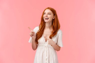 Karizmatik, sevimli Avrupalı, beyaz moda elbiseli, komik bir şey gösteren, sol üst köşeyi işaret eden ve bakan, yüksek sesle gülen, mutluluk ve neşe ifade eden kızıl saçlı kadın.
