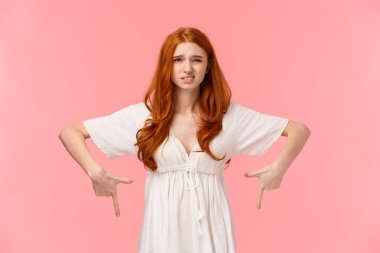Well I dont know. Waist-up portrait indecisive and unsure, slightly displeased cute redhead woman in white dress, pointing down, cringe as express dislike, disappointed with gift, pink background