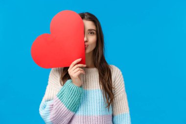 Romantic gesture, love and valentines day concept. Dreamy attractive girlfriend in winter sweater, cover half face with red heart, folding lips in kiss, standing blue background showing sympathy
