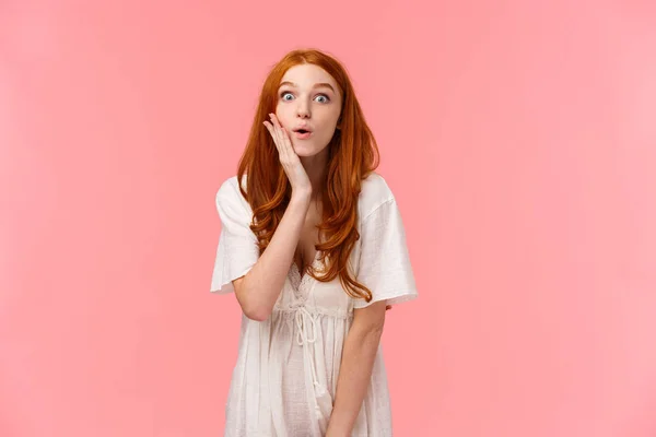 Curious, fascinated cute redhead european woman in white dress, touch cheek and sighing, gasping astonished, look camera intrigued and amused, seeing something incredible, pink background — Stock Photo, Image