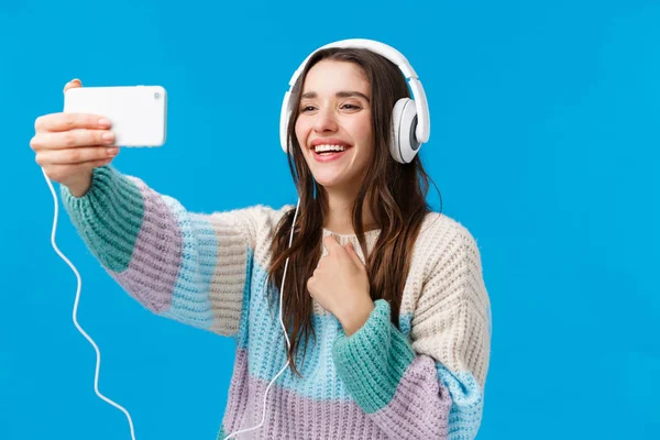 Encantador y lindo novio de contacto novia a través de Internet, videollamadas en línea, la celebración de brazo extendido smartphone, el uso de suéter de invierno, auriculares grandes, tomando selfie y sonriendo, fondo azul — Foto de Stock