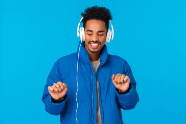 Guy catching positive vibes, feeling groovie and satisfied with new headphones. Cheerful african american guy wearing winter padded jacket, bought earphones holidays sale, dancing and listen music