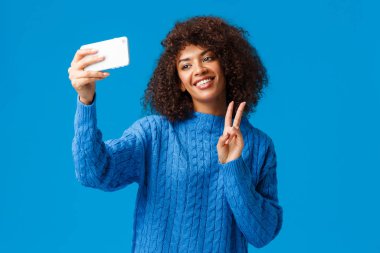 Neşeli arkadaş canlısı ve sevimli Afro-Amerikalı kız öğrenci fotoğraf çekiyor. Yeni akıllı telefon uygulamasına filtre uyguluyor, selfie çekiyor, başını eğiyor, gülümsüyor, barış jesti yapıyor, mavi arka plan.
