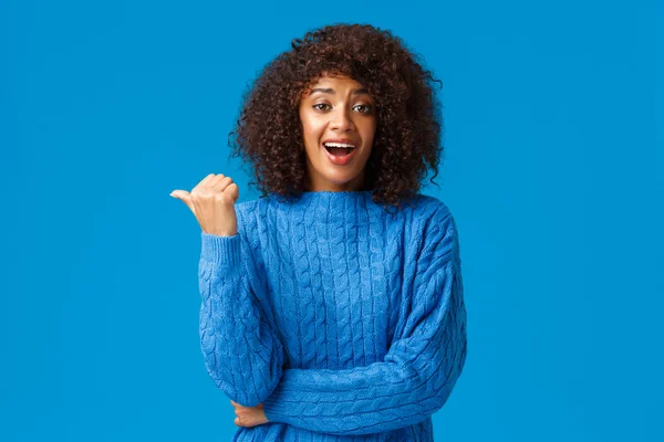 Excited happy pretty african-american woman with afro haircut in winter sweater, pointing thumb left and laughing as discuss funny moment, talking casually during party, blue background — 스톡 사진