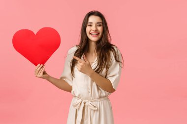 Ruh eşi aranıyor. Neşeli, güzel, gülen bir kız, büyük kırmızı bir kalp taşıyor, sevgililer günü kartonu ve kamerayı işaret ediyor, sevgi ve sevgi gösterisi istiyor, pembe arka plan.