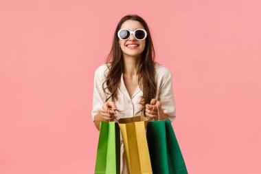 Kadın alışverişe gitti, yeni kıyafetler aldı, tatil sezonu için özel indirimler aldı, mağaza çantaları ve gülümseyen kameralarla durdu, güneş gözlüğü taktı, pembe arka plan heyecanlıydı.