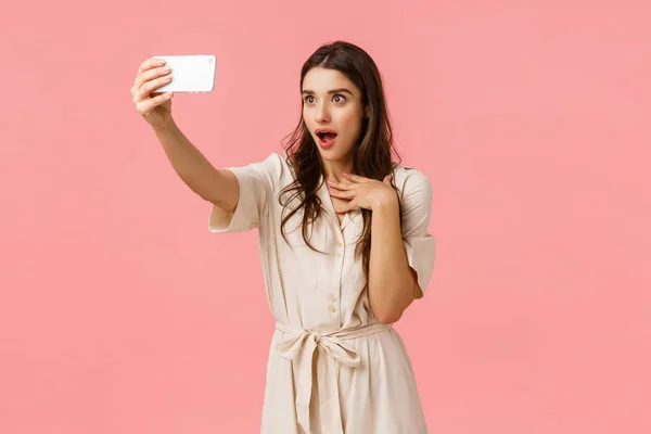 Chica haciendo fascinado y sorprendido expresión como hablar con la novia en la videollamada telefónica. Encantador morena hembra en vestido, tomando selfie, jadeando boca abierta divertido, de pie fondo rosa —  Fotos de Stock