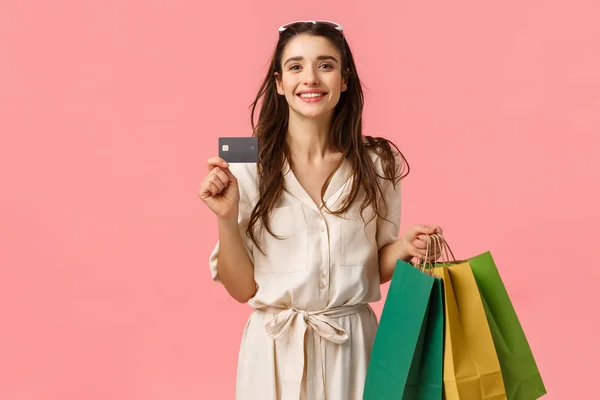 Glad kaukasiska brunett kvinna i snygg klänning, bära shoppingväskor och kreditkort, betala allt med betalkort, har särskild rabatt, med bankdepositionen, stående rosa bakgrund — Stockfoto