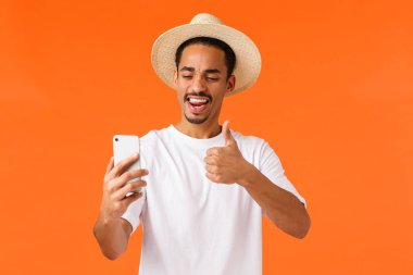 Çekici, mutlu, genç, Afro-Amerikan erkek turist tatilde, akıllı telefondan selfie çekiyor ve onaylama, onaylama, yüzsüzlük, sevinç ifadesi, turuncu arka plan