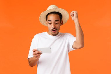 Rahatlamış ve mutlu, zafer kutlayan Afro-Amerikan genç adam, oyunda sert seviyeyi geçti, cep telefonu ekranını tutuyor, tezahürat içinde elini kaldırıyor, turuncu arka plan