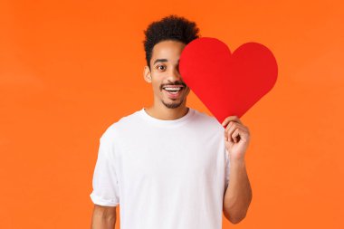 Sevgililer günü, ilişki ve takdir konsepti. Çekici, romantik erkek arkadaş, Afro-Amerikan adam, kalp kartını yüzüne yakın tutuyor ve mutlu bir şekilde gülümsüyor, gerçek aşkı arıyor, turuncu arka plan