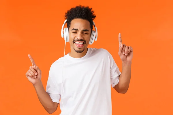 Unbekümmert fröhlicher und erleichterter afrikanisch-amerikanischer gutaussehender Hipster, der mit Kopfhörern Musik hört, im Rhythmus tanzt und Hände schüttelt, die Augen schließt und mit Kopfhörern mitsingt, orangefarbener Hintergrund — Stockfoto