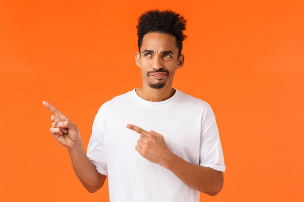 Skeptical and reluctant disappointed african-american man, afro haircut and moustache, pointing looking upper left corner displeased, smirk unsatisfied and judgemental, orange background — Stock Photo, Image