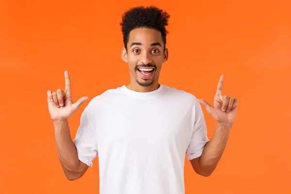 Excited and intrigued, enthusiastic african-american male in t-shirt, with moustache afro haircut, pointing fingers up, smiling amused, showing perfect product, shopping center, orange background — Stock Photo, Image