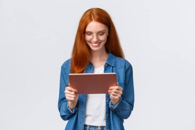 Belden yukarı portre oldukça mutlu kızıl saçlı kız öğrenci, çalışıyor, tasarım projesi üzerinde çalışıyor, gözlük takıyor, dijital tablet tutuyor ve tatmin olmuş görünüyor.