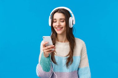 Teknoloji, milenyum ve yaşam tarzı kavramı. Kaygısız tatlı esmer kız öğrenci kulaklık taktı, akıllı telefon seçimi şarkısı taktı ve gülümsedi, mavi arka planda durdu çalışma için çalma listesi yaptı.